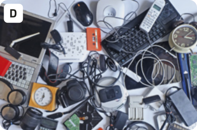 D. Fotografia de muitos aparelhos eletrônicos amontoados em uma mesa. Há máquinas fotográficas, teclados, relógios analógicos, 'mouses', carregadores, um telefone, um computador, e muitos fios.