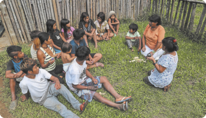 Fotografia de crianças e adolescentes indígenas sentados na grama, ao lado de uma cerca de madeira. Eles estão próximos uns dos outros e escutando atentamente a uma mulher adulta indígena, que está sentada sobre os calcanhares.