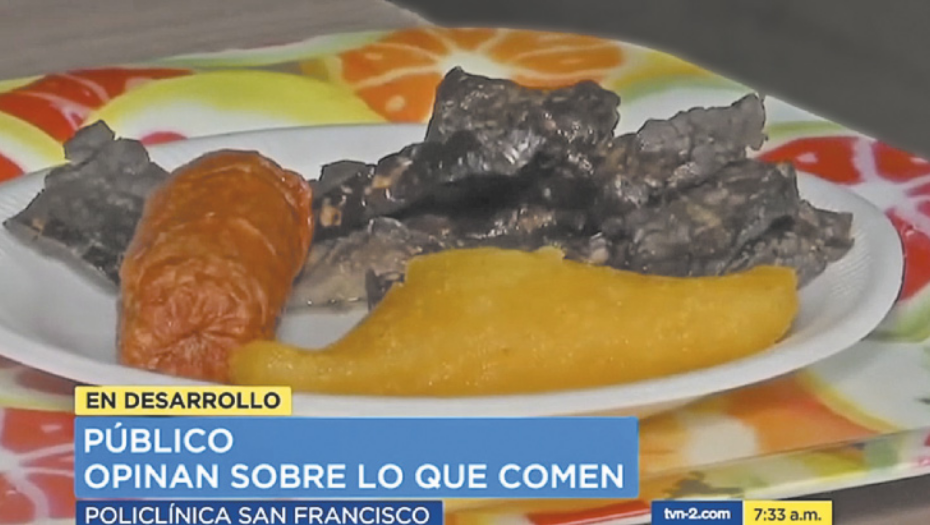 Imagem de uma reportagem de televisão, contendo um prato de comida. O prato contém dois tipos de carne e um pedaço de legume. O prato está acima de uma bandeja estampada com laranjas cortadas. Abaixo da foto está escrito: 'En desarollo. Público opinan sobre lo que comen. Policlínica San Francisco'.