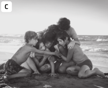 C. Fotografia em preto e branco de crianças se abraçando em uma praia. As crianças estão sentadas na areia, e têm uma expressão triste, com tom de consolo. Elas estão usando trajes de banhos. Ao fundo está o mar com ondas.