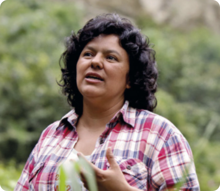 Fotografia de Berta Cáceres. Ela tem cabelos cacheados na altura dos ombros, usa uma camisa xadrez e está gesticulando com uma das mãos, com a boca aberta, como se estivesse falando. Ao fundo, desfocado, há vegetação.