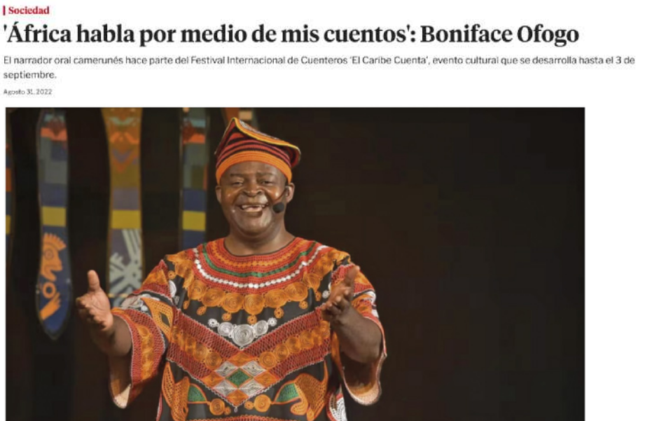 Imagem de uma página de notícias na internet. A notícia faz parte da seção de 'Sociedad' do site, e a manchete é a seguinte: 'África habla por medio de mis cuentos': Boniface Ofogo'. Abaixo da manchete, em letras menores, está escrito: 'El narrador oral camerunés hace parte del Festival Internacional de Cuenteros 'El Caribe Cuenta', evento cultural que desarolla hasta el 3 de septiembre'. A notícia é de 31 de agosto de 2022. Há uma fotografia de Boniface Ofogo, que é negro, e está usando um traje africano muito colorido e com figuras geométricas, além de um chapéu. Ele está sorrindo, e tem um microfone pequeno preso ao rosto. Seus braços estão estendidos com as palmas das mãos para cima. Ao fundo, há três faixas com figuras representando a cultura africana.