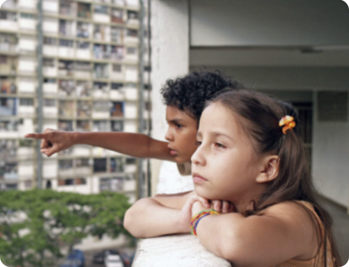 Fotografia de duas crianças olhando a vista da sacada de um prédio. Há um menino negro de cabelos cacheados apontando algo à distância. Ao seu lado está uma menina branca de cabelos lisos e claros, presos com maria chiquinhas. A menina tem os braços apoiados no muro da sacada e está olhando para onde o menino aponta com uma expressão tranquila. Ao fundo, há outro prédio com muitas janelas, e árvores.