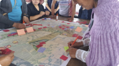 Fotografia de pessoas em volta de uma mesa onde há um grande mapa. O mapa está coberto com diversos papéis do tipo 'post it' coloridos, e uma das pessoas está escrevendo algo em um destes papéis.