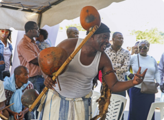 Fotografia de um homem negro segurando um instrumento musical, falando e gesticulando para um grupo de pessoas. O homem tem uma expressão alegre, e está usando um gorro, regata, calças largas e um bracelete. Com uma das mãos ele gesticula, e com a outra segura um instrumento musical composto por uma vara de madeira com três grandes cabaças amarradas. Ao fundo, há pessoas negras observando a cena. Todos estão debaixo de uma tenda ao ar livre, e na parte direita da fotografia há uma paisagem ensolarada.
