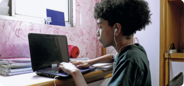Fotografia de um jovem usando um computador. Ele está sentado na frente de uma mesa de estudos, usa fones de ouvido com fio, e uma camiseta. Em cima da mesa de estudos, há livros, caderno e estojo. À frente do jovem, há uma janela que ilumina o ambiente.