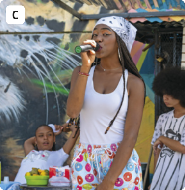 C. Fotografia de uma jovem cantando rap e segurando um microfone perto da boca. Ela tem o cabelo trançado e usa uma bandana, regata e bermuda colorida. A fundo há outros dois jovens, uma sentada e o outro em pé, assistindo à apresentação. As paredes do ambiente são pintadas com ilustrações coloridas.