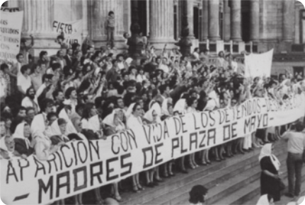 Fotografia em preto e branco da manifestação das avós da Praça de Maio. Há diversas pessoas, em pé nas escadas de entrada da Casa Rosada, a sede do governo argentino. Elas seguram um cartaz com os dizeres: 'Aparición con vida de los detenidos', e 'Madres de Plaza de Mayo'. Ao fundo, o edifício é composto por diversas pilastras cilíndricas.