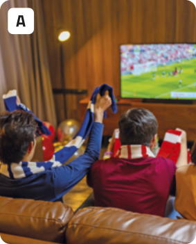A. Fotografia de dois jovens vistos de costas. Eles estão sentados em um sofá de couro assistindo a uma partida de futebol em uma televisão. Os jovens estão usando roupas e cachecóis listrados de cores diferentes, e estão torcendo e vibrando com os braços para cima. Ao fundo, há a televisão em cima de um móvel de madeira, uma luminária alta e longas cortinas à esquerda.