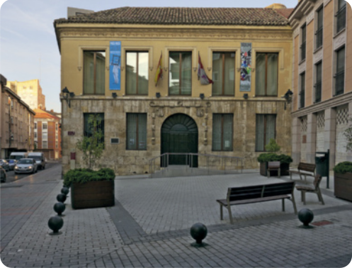Fotografia do Museu de Palência. Trata-se de uma construção de dois andares com uma entrada em forma de arco e diversas janelas retangulares compridas, dispostas lado a lado. Há cartazes e bandeiras entre algumas das janelas, e corrimões na frente da porta. Na frente do prédio, há uma praça com chão de paralelepípedos, onde há alguns bancos, lixeiras, canteiros com plantas, e estruturas compostas por esferas próximas ao chão. Ao fundo, há uma rua com carros e edifícios. O céu está azul com algumas nuvens esparsas.