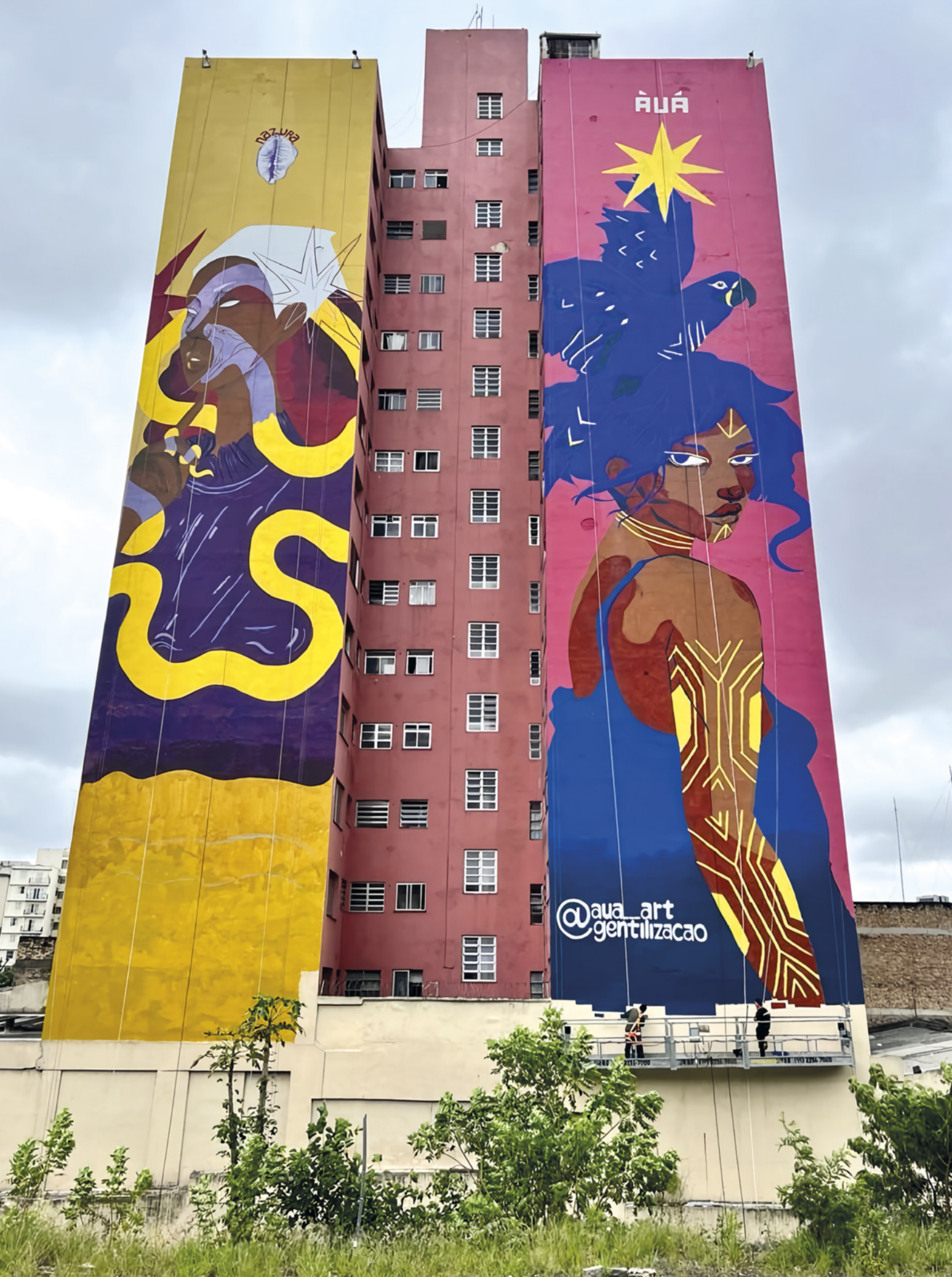 Fotografia de um prédio alto com murais coloridos em um contexto urbano. No centro do prédio há muitas janelas retangulares dispostas verticalmente. Na fachada da esquerda, há a ilustração de uma mulher negra usando um pano envolvendo seus cabelos. Em volta da mulher há uma serpente cuja boca se abre próximo à boca da mulher. Na fachada da direita, há a ilustração em vista lateral de uma mulher negra com cabelos que se transformam em uma arara-azul. O corpo da mulher está pintado em tons alternados de marrom escuro e marrom claro. Seu rosto, pescoço e braço estão pintados com linhas e setas, e ela está usando uma regata. Ao redor da base do prédio, há grama e arbustos. Ao fundo, há outros prédios mais baixos e o céu nublado.