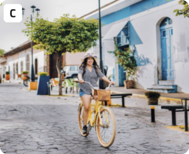 C. Fotografia de uma jovem andando de bicicleta em um ambiente urbano. A jovem está sorrindo e usando camisa de botões, bermuda jeans, chapéu e tênis. Ao fundo, a paisagem é uma rua de blocos de pedra com bancos, postes de iluminação, plantas, e um prédio baixo com portas altas.