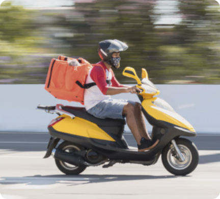 Fotografia de um entregador de comida dirigindo uma moto. O homem que dirige a moto está usando bermuda, tênis, camiseta com as mangas até os cotovelos, e um capacete com o visor levantado. Nas costas, ele carrega uma sacola grande e quadrada, que serve para guardar os pedidos. A moto é do tipo 'scooter' de pequeno porte, e aparenta estar em movimento, pois o cenário de fundo está desfocado.
