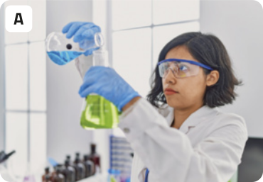 A. Fotografia de uma jovem fazendo um experimento científico. A jovem está usando óculos e luvas de segurança, além de um jaleco branco. Com as mãos, ela despeja um líquido azul contido em um béquer, dentro de outro béquer, que contém um líquido verde. Ao fundo, o cenário é de um laboratório, todo branco, com alguns frascos organizados em uma bancada, e janelas grandes.
