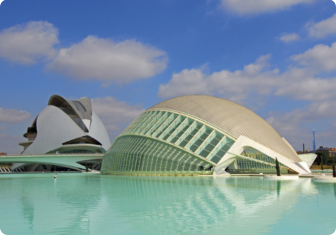 Fotografia de uma construção com arquitetura moderna, acima de uma piscina de água rasa. À esquerda, há um edifício formados por duas placas metálicas côncavas juntas, assemelhando-se a uma concha. À direita, há outro edifício com formato ovalado, formado por uma parte sólida, e outra com várias estruturas metálicas verticais separadas por vidraças. Ao fundo, o céu está azul com algumas nuvens.