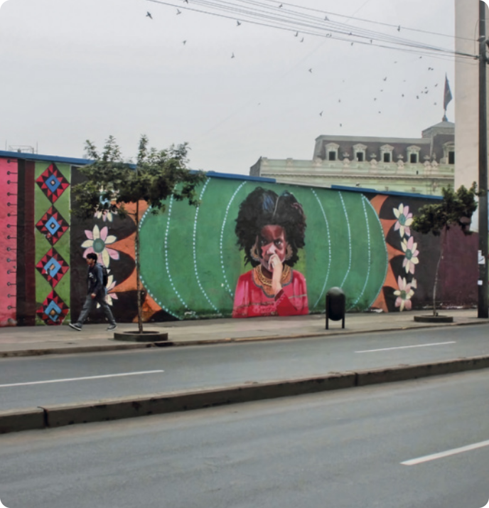 Fotografia de um mural de rua. O mural representa uma pessoa negra com cabelo 'black power', usando brincos e roupas africanas. De ambos os lados da pessoa, estão representadas flores e formas geométricas de losangos e triângulos. A arte está localizada na calçada de uma rua vazia, que possui uma lixeira e árvores. Há uma pessoa caminhando na calçada e atrás do muro, ao fundo, há uma construção.