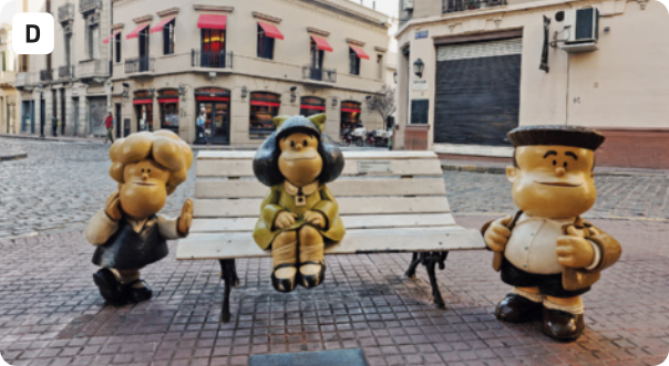 D. Fotografia de uma praça com esculturas de três personagens das histórias em quadrinhos da Mafalda. A escultura que representa a Mafalda está sentada em um banco de madeira no centro da foto. À sua esquerda, apoiada no banco, está Susanita. À direita, em pé, está Manolito. Ao fundo, há prédios antigos com toldos sobre as janelas. O chão é de lajotas quadradas.