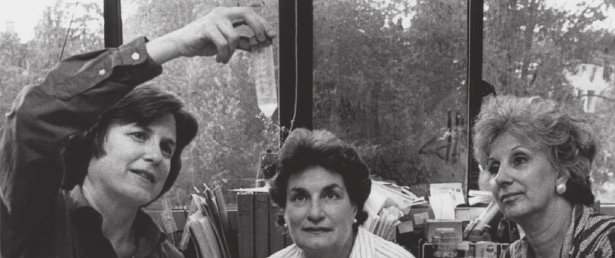 Fotografia em preto e branco de três mulheres. À esquerda, a geneticista Mary-Claire King segura um tubo contendo um líquido, que mostra para duas senhoras à sua direita. Mary-Claire é branca e tem os cabelos escuros, lisos e curtos. Ela está usando uma camisa. Ao seu lado, as senhoras são brancas e têm os cabelos curtos. Uma é morena, e a outra loira, e ambas olham para o tubo. Ao fundo, há uma estante com pastas e papéis, e uma janela ampla com vista para vegetação.