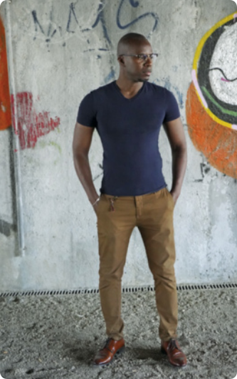 Fotografia de Edjanga Jones Ndjoli, em pé com as mãos nos bolsos. Ele é negro, careca, está usando óculos de grau, camiseta, calça e sapato social. Sua expressão é séria. Ao fundo há uma parede com alguns grafites.