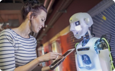 Fotografia de uma mulher e um robô com forma humana. A mulher está digitando algo em um tablet, com o robô à sua frente. O robô tem uma aparência humanoide, com rosto e tronco, porém sem os braços. No lugar de onde sairia um dos braços e no centro do peito do robô, há uma variedade de fios e engrenagens. O robô está iluminado no rosto e tronco. A mulher tem os cabelos presos e usa uma camiseta.