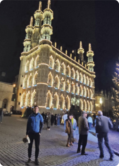 Fotografia de Aimar posando na praça Grote Markt, na Bélgica. Aimar é um jovem branco de cabelos curtos e escuros, e está usando jaqueta e calça. Ele está na frente de uma construção com diversas torres e janelas iluminadas. Há outras pessoas passeando pela praça, e o céu está escuro.