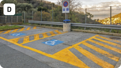 D. Fotografia de uma vaga para pessoas com deficiência em um estacionamento. Há listras pintadas de amarelo em volta da vaga, que contém uma figura de uma pessoa usando cadeira de rodas pintada no chão. Há uma placa e vegetação ao fundo.