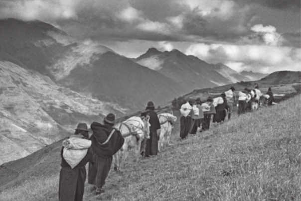 Fotografia em preto e branco de um grupo de pessoas caminhando em fila indiana por uma encosta gramada e íngreme. As pessoas estão usando roupas escuras. Algumas estão levando cavalos, que carregam fardos em seus lombos, e outras estão carregando sacos nas costas. Ao fundo, há uma cadeia de montanhas e um céu cheio de nuvens.