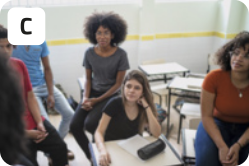 C. Fotografia de jovens em uma sala de aula. Uma jovem está sentada em uma carteira, enquanto outros jovens estão em pé apoiados nas carteiras com expressões interessadas.