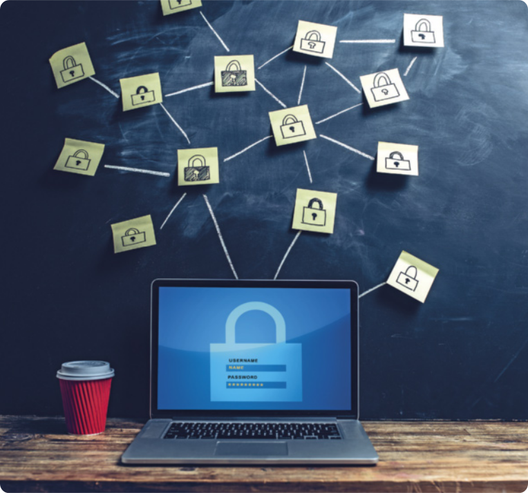 Fotografia de um computador em uma mesa de madeira. Na tela do computador, há a ilustração de um cadeado com espaço para 'login' e senha. Ao lado do computador há um copo de café com tampa. Na parede atrás da tela do computador, há muitos papéis do tipo 'post-it', com desenhos de cadeados, colados na parede e interligados por linhas, formando uma rede.