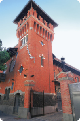 Fotografia da fachada de um museu. A construção é feita de tijolos e possui uma torre alta e quadrada com janelas de diferentes tamanhos e formatos em cada andar. No topo da torre, há um telhado, logo acima de uma sacada com colunas adornadas, dando um aspecto medieval à construção. Perto do chão há um portão de metal aberto, cercado por duas colunas de tijolos. Ao fundo, o céu está claro, sem nuvens.