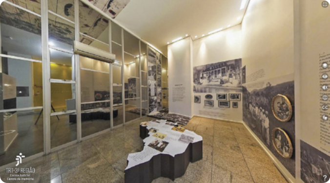 Fotografia de um ambiente em uma exposição de museu. O chão é de pedra polida e à esquerda há uma parede coberta por espelhos retangulares, com um aparelho de ar condicionado posicionado no alto. No centro do espaço, há uma estrutura em 3D com o formato do mapa do Brasil, com informações e fotografias ilustrando a parte superior. Na parede à direita, há diversas fotografias em preto e branco, algumas maiores, outras menores e dispostas em molduras circulares. Há também muitas informações escritas.