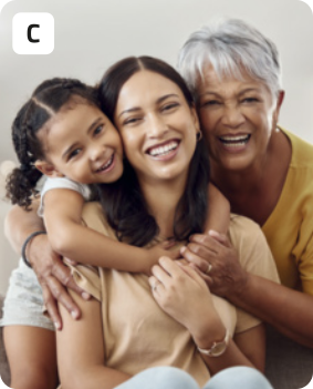 C. Fotografia de duas mulheres adultas e uma criança sorrindo abraçadas. No centro da fotografia está uma mulher jovem de cabelos lisos e escuros, sorrindo e usando uma camiseta. Abraçando a mulher, estão uma menina e uma mulher mais velha. A menina tem os cabelos presos com maria-chiquinhas, e a mulher mais velha está usando camiseta amarela e tem os cabelos grisalhos cortados curtos.