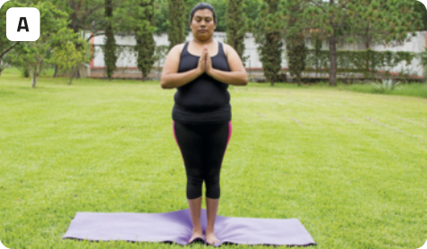 A. Fotografia de uma mulher em pé, em uma postura meditativa. A mulher tem os cabelos presos e está usando roupas justas, de ginástica. As mãos da mulher estão juntas, como em oração, ela está com os olhos fechados e posicionada acima de um tapete. O cenário é de um parque, com grama baixa e árvores ao fundo.