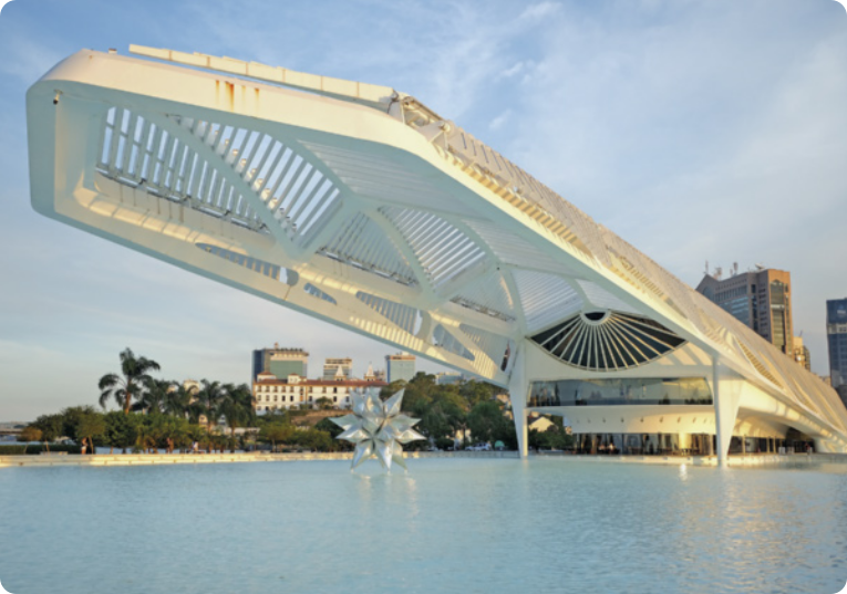Fotografia do Museu do Amanhã. O cenário é composto por uma construção de arquitetura moderna, contendo um edifício principal com grandes arcos de metal brancos que se elevam e se curvam, formando uma cobertura que se projeta sobre um grande espelho de água. Abaixo desta estrutura e no meio do espelho de água, há uma escultura com muitas extremidades pontudas. Na paisagem ao fundo há várias árvores, coqueiros e edifícios altos, e o céu está levemente nublado.