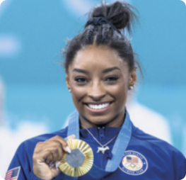 Fotografia de Simone Biles. Ela é negra e está com os cabelos presos em um coque no alto da cabeça. Ela está sorrindo, segurando uma medalha de ouro que adorna seu pescoço com uma das mãos, e usando uma jaqueta azul fechada com o símbolo dos Estados Unidos.