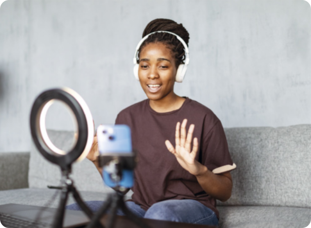 Fotografia de uma jovem sentada em um sofá acenando para a câmera de um celular apoiado em um tripé. A jovem está usando cabelo preso, fones de ouvido, camiseta e calça jeans. À sua frente há um arco de iluminação comumente utilizado por criadores de conteúdo na internet.