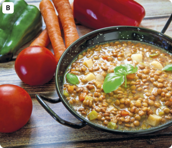 B. Fotografia de uma panela de ferro contendo um cozido de lentilhas. A comida está enfeitada com uma folhinha de tempero, e ao lado há tomates, cenouras e pimentões. A panela está disposta em cima de uma mesa de madeira.
