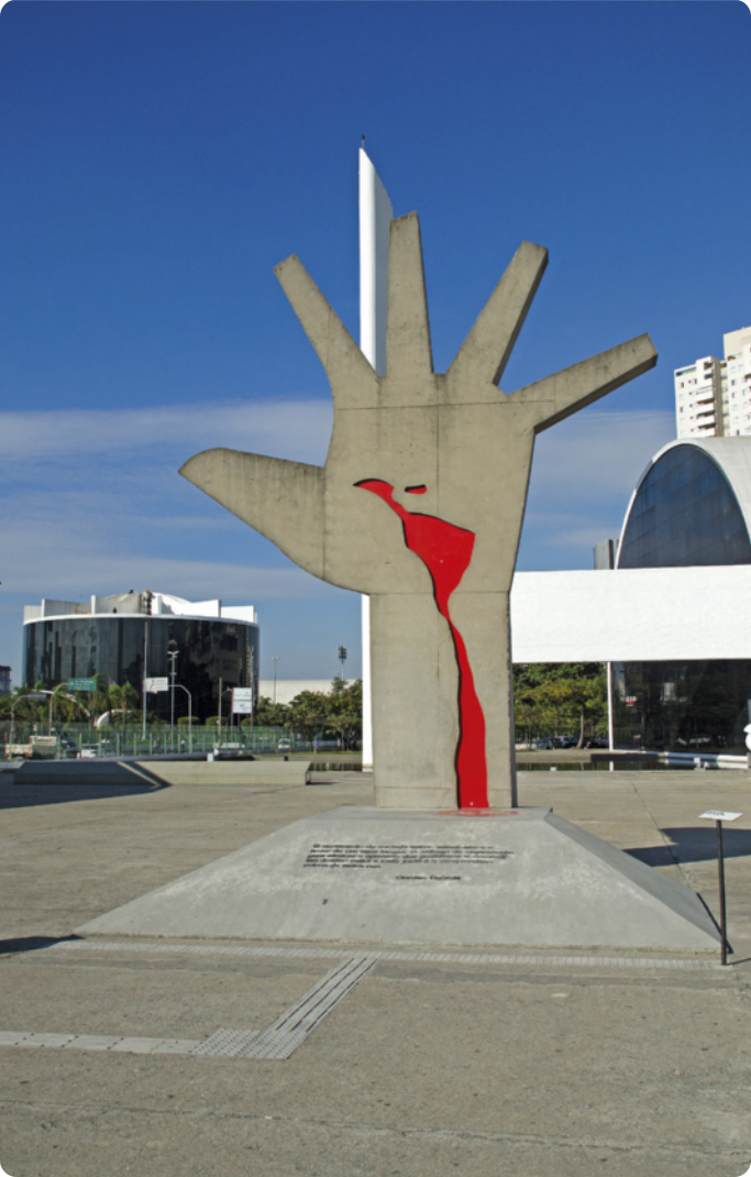 Fotografia do Monumento à América Latina. O monumento é uma escultura que representa a mão aberta de uma pessoa, com o mapa da América Latina desenhado em vermelho no centro da palma até o pulso. Na base da escultura há informações escritas em uma placa. Ao fundo, há ruas, carros, e edifícios.