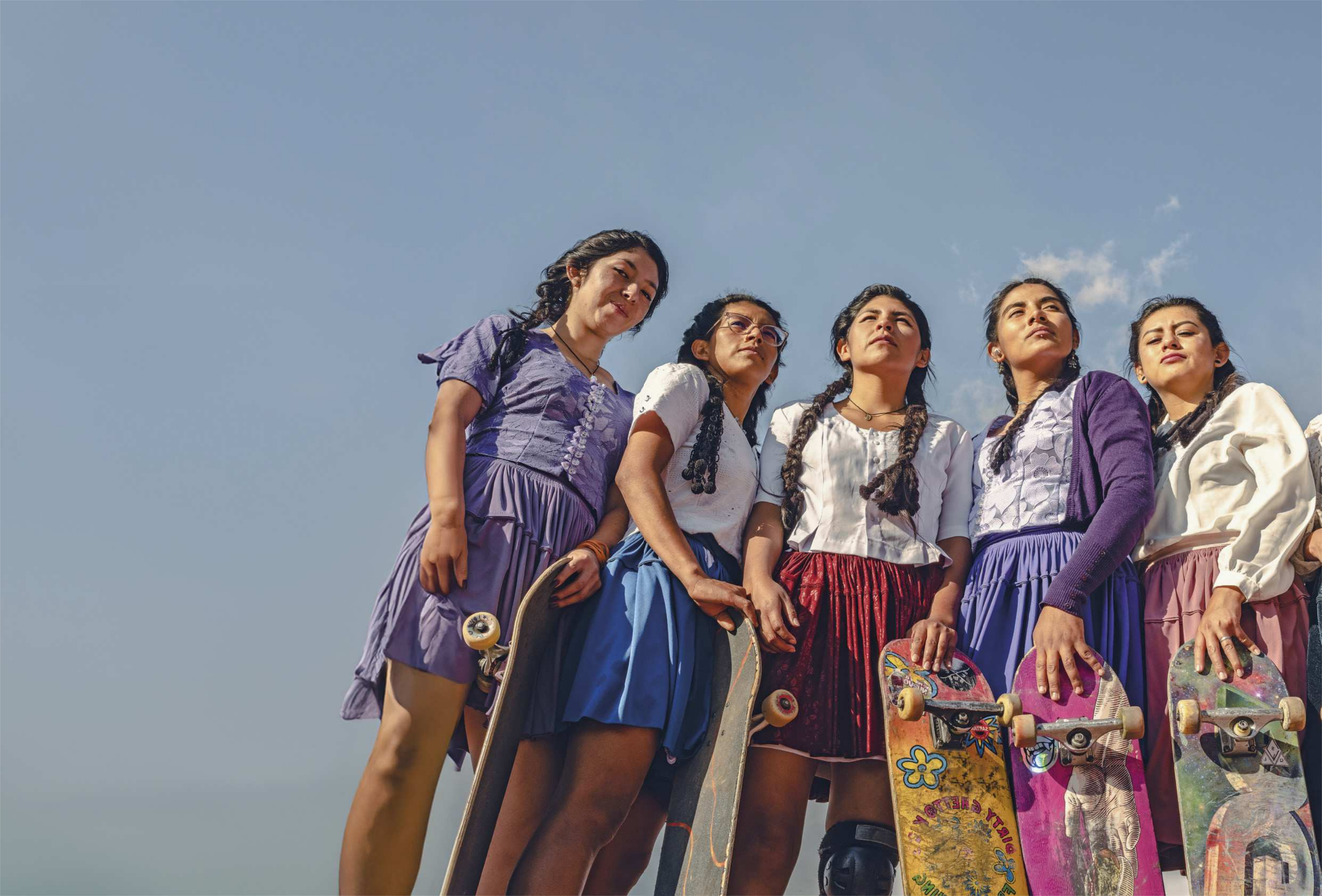 Fotografia de abertura de Unidade. Há cinco jovens de traços indígenas olhando para a frente, vistas de baixo para cima. Elas estão usando roupas tradicionais da sua cultura, que consistem em saias e blusas bordadas, e estão segurando skates. Uma delas usa um casaco em cima da blusa. A expressão das jovens é séria e determinada. Ao fundo, o céu está claro e azul.