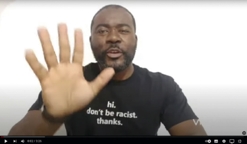 Imagem de um vídeo do canal de 'cuentos africanos Potopoto'. Há um homem acenando aos espectadores com uma expressão amigável. Ele é negro, ele tem cabelo curto, barba e bigode, e está usando uma camiseta com os dizeres: 'hi. don't be racist. thanks'. Na parte inferior da imagem, está a barra de reprodução do vídeo, com botões de 'play', acelerar, volume, legenda, configurações e modo de tela cheia.