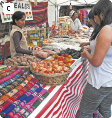 C. Fotografia de duas mulheres comprando produtos em uma tenda da feira. As duas mulheres estão olhando para alguns legumes dispostos em uma bancada com toalha listrada. Há pimentão, batata e milho organizados em cestos, alguns tecidos coloridos, entre outros produtos. Atrás da bancada, está uma vendedora. Ao fundo, há outra vendedora.