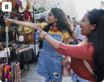 B. Fotografia de duas jovens olhando e apontando para algo em uma feira de rua. Uma das jovens está usando fones de ouvido ao redor do pescoço, camiseta, macacão jeans e uma bolsa em um dos ombros. A outra está usando camiseta de mangas até os cotovelos e mochila. Ambas estão sorrindo e tem os cabelos compridos e escuros, soltos. Ao fundo, há uma tenda da feira com diversos produtos expostos, como chapéus e colares.
