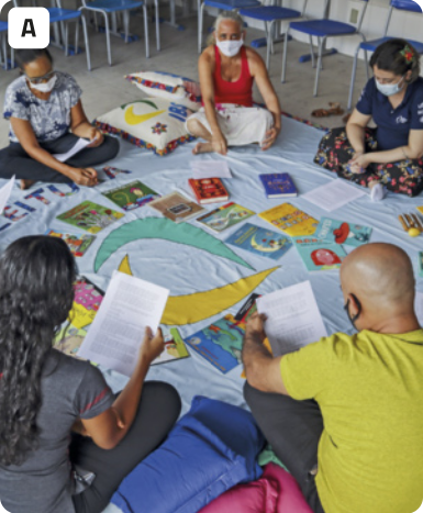 A. Fotografia de diversas pessoas sentadas no chão em uma roda. As pessoas estão sentadas com as pernas cruzadas em cima de um pano onde estão dispostos vários livros infantis. Elas estão com máscaras cirúrgicas cobrindo seus narizes e bocas. Três pessoas seguram e olham para uma folha de papel sulfite com um texto. Há almofadas ao lado de uma das pessoas. Ao fundo, há diversas cadeiras escolares.