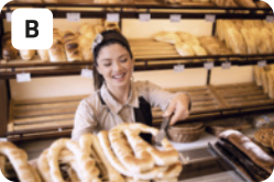 B. Fotografia de uma mulher de uniforme sorrindo e pegando um 'pretzel' com um pegador, atrás de um balcão. Ao fundo há várias prateleiras com diferentes tipos de pães.