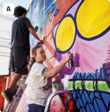 A. Fotografia de dois jovens grafitando um grande painel. Há um jovem em cima de uma escada pintando o painel com uma lata de tinta, enquanto se segura à escada com a outra mão. Ele usa camiseta, bermuda e tênis. Mais abaixo, também em uma escada, uma jovem está pintando outra parte do painel. Ela usa camiseta e calça. O painel tem figuras coloridas.