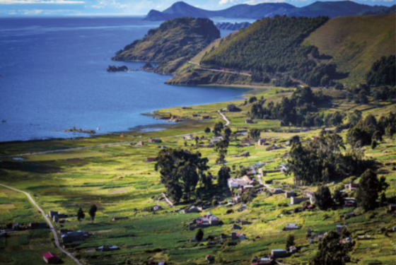 Fotografia de uma paisagem composta com um relevo coberto com vegetação baixa, algumas árvores, construções e estradas, com montanhas e um grande lago ao fundo.