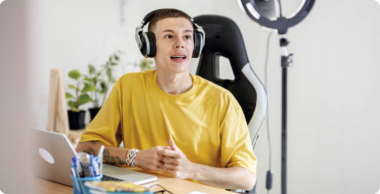 Fotografia de um jovem gravando um vlog. O jovem está falando, sentado em uma cadeira, e apoiando os braços em uma mesa com um computador à sua frente. Ele tem cabelos curtos e escuros e está usando camiseta e fones de ouvido. Ao seu lado há um arco de iluminação, comumente utilizado por criadores de conteúdo na internet. Ao fundo, há alguns vasos de plantas.