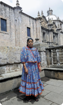 Fotografia de Lorena Ramírez em pé na frente de uma construção feita de blocos de pedra, com torres, janelas e muretas baixas. Lorena tem traços indígenas e está usando um vestido florido, que possui uma saia rodada e mangas largas que vão até os cotovelos, uma bolsa com a alça atravessada no peito, e sandálias. Ela está usando os cabelos presos e tem uma expressão séria.