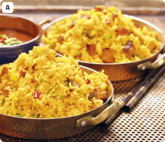 A. Fotografia de duas panelas contendo arroz com frango. O arroz tem coloração amarela, há pedaços de frango e de pimentão vermelho. As panelas são de ferro e estão dispostas em um tipo de chapa com muitos buracos pequenos e circulares. Ao lado das panelas, há dois palitos ornamentados que aparentam 'hashis' da culinária oriental, e ao fundo há mais uma tigela com outro tipo de comida.