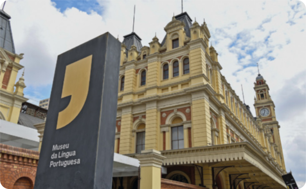 Fotografia do Museu da Língua Portuguesa. Há um monumento retangular de pedra escura com um grande símbolo de vírgula, acompanhado da inscrição 'Museu da Língua Portuguesa'. Ao fundo, está a fachada da Estação da Luz, que abriga o Museu, com torres e janelas. Ao fundo, o céu está cheio de nuvens.